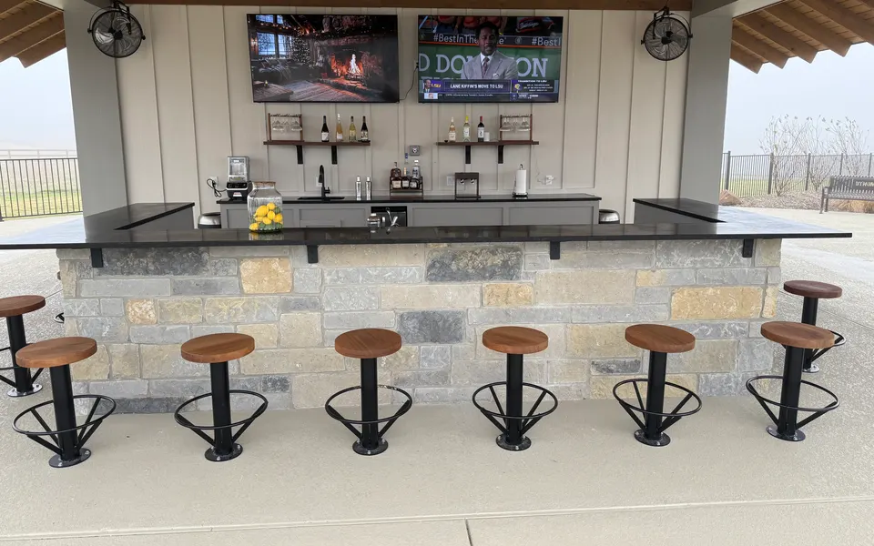 Covered outdoor bar with stone base and black countertop, seven fixed stools, two TVs and ceiling fans