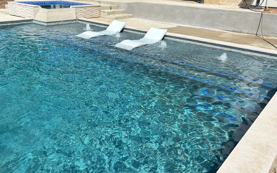 Two in-water loungers on a tanning ledge with bubblers, raised blue-tile spa with sheer descent behind